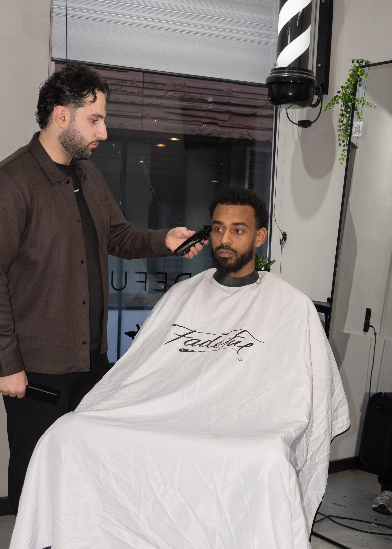 Een barber trimt de baard van een klant in een witte Fadeful-kaap, met de barberpaal en planten achter hen