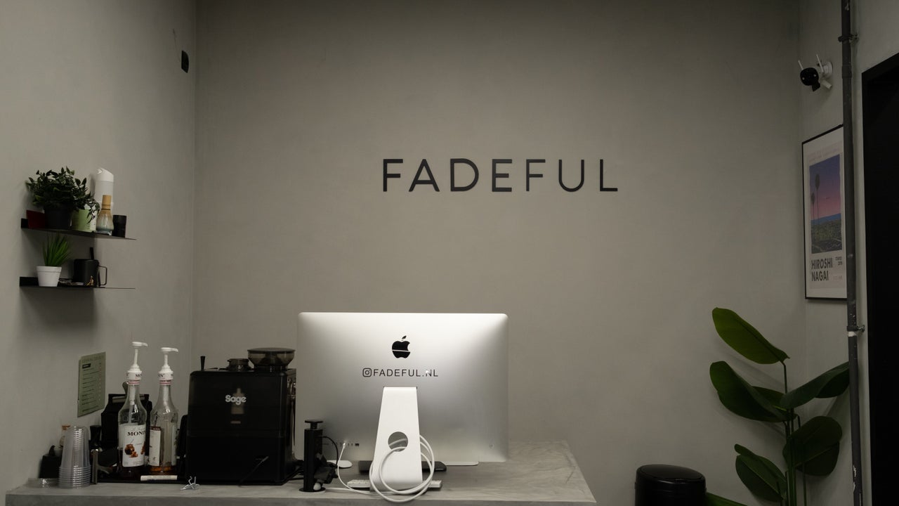 De receptie van Fadeful Barbershop met de letters FADEFUL op de betonwand, een iMac en planten